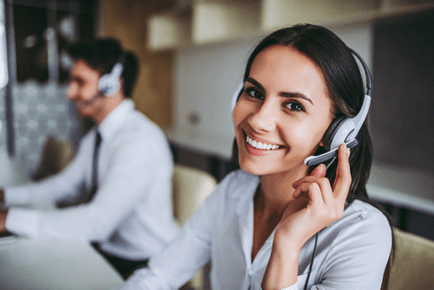 A smiling person wearing a headset is in the foreground; another person with a headset is in the blurred background, suggesting a professional office setting.