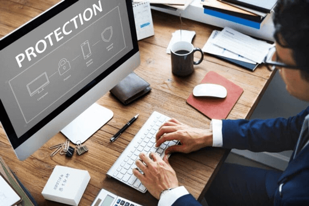 A person is working at a desk with a computer screen displaying the word "PROTECTION" alongside icons of a lock, computer, house, and shield.